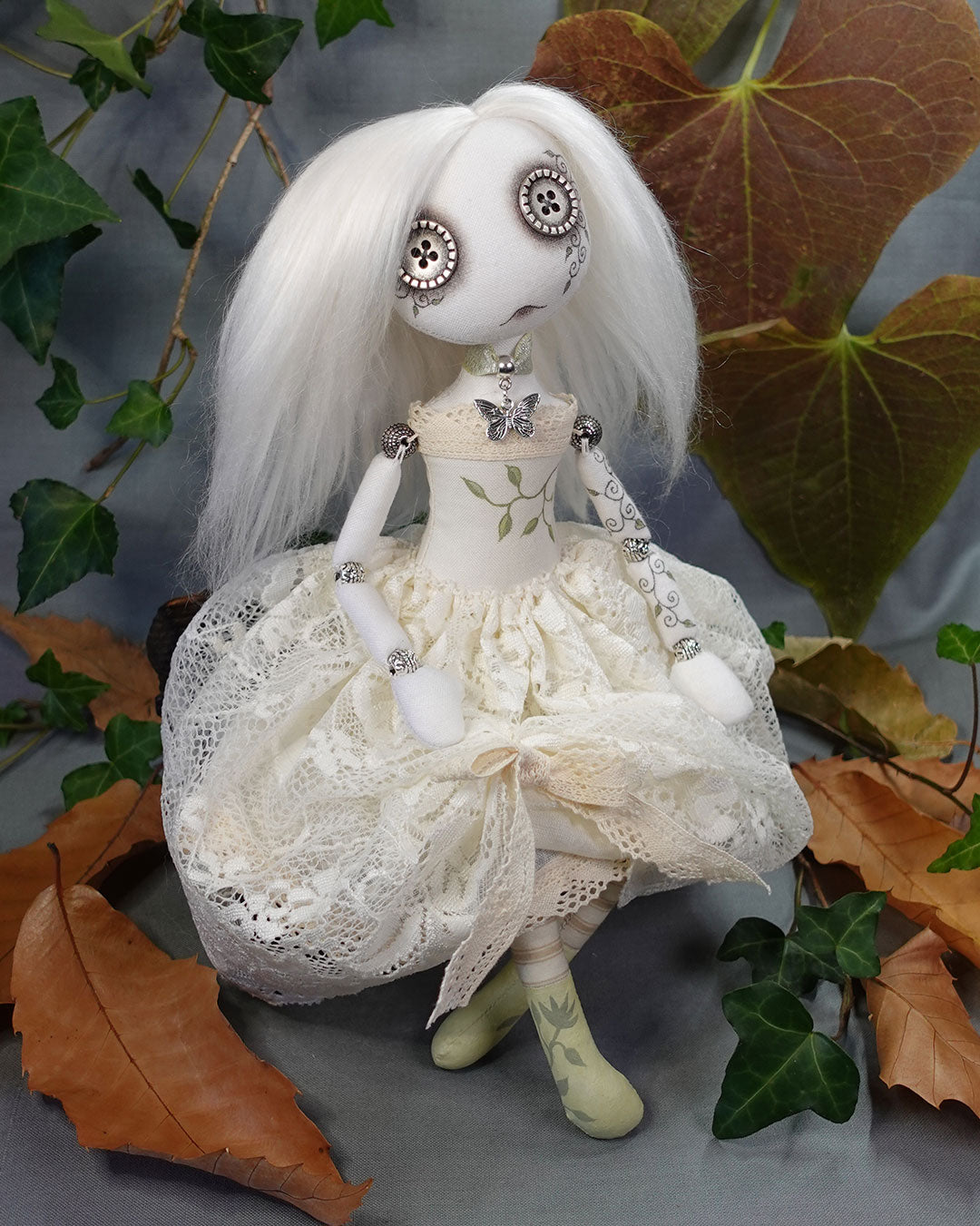 Textile art doll with white hair and detailed clothing among leaves