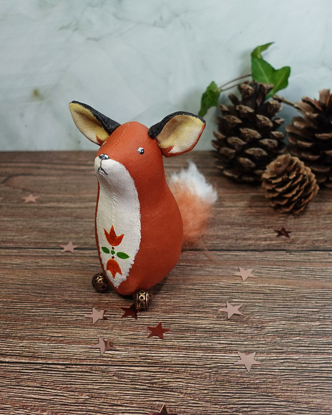 Handmade fox figurine with floral design on a wooden surface with pine cones and leaves.