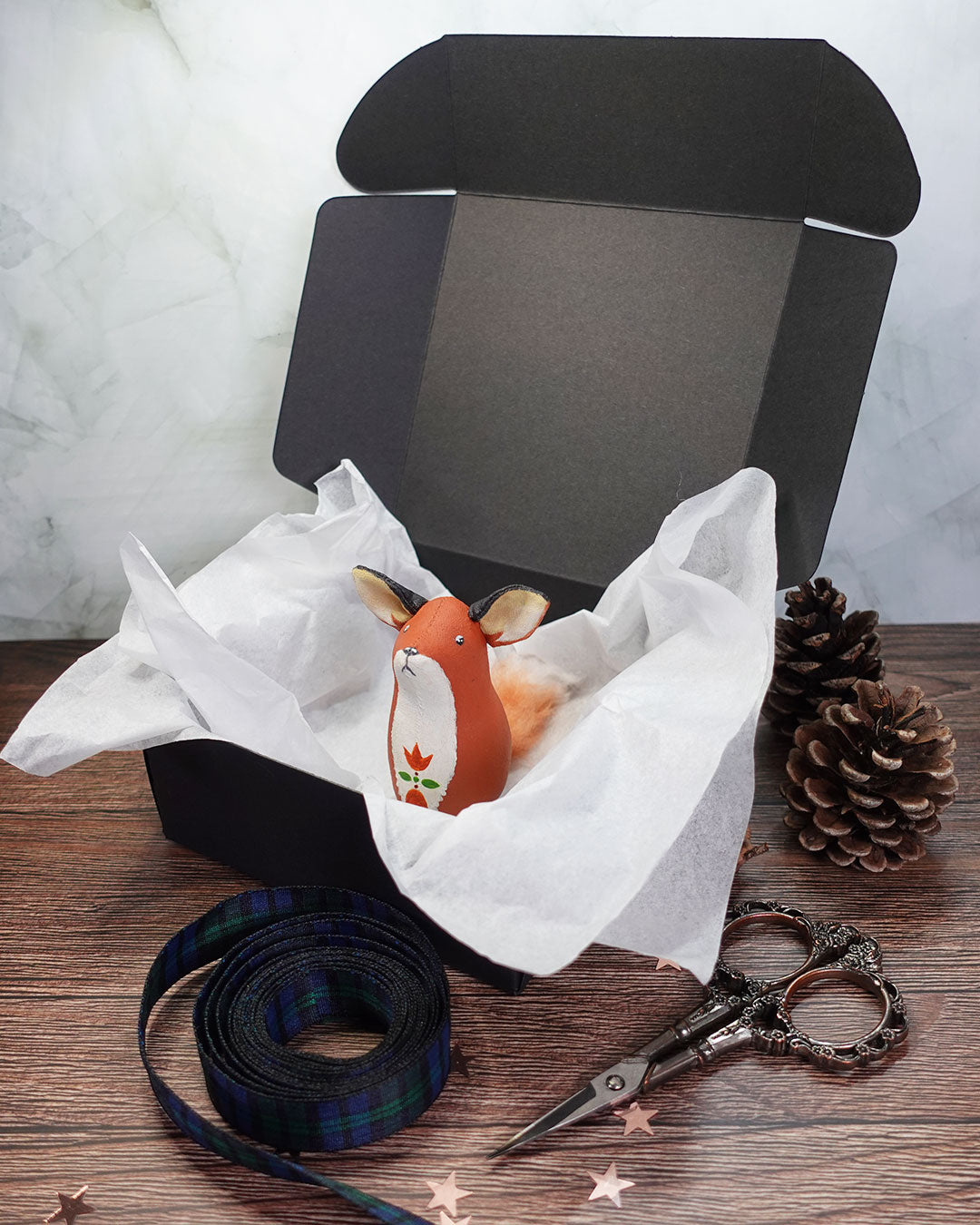 Small fox figurine in a black box with white tissue paper, surrounded by decorative items on a wooden surface.