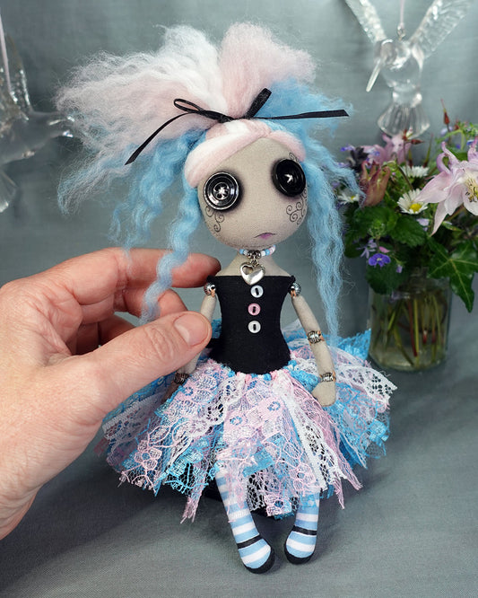a button eyed cloth art doll in trans flag colours, shown with artist's hand for scale