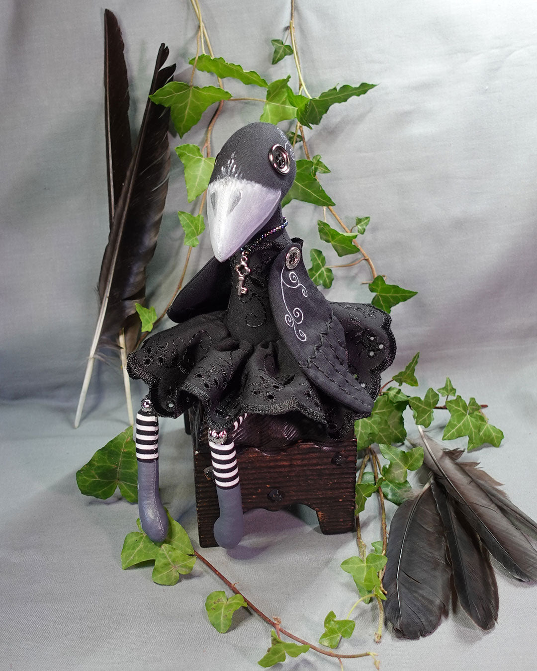 Button eyed crow art doll with painted beak, black dress, and striped legs on a grey background with feathers and ivy.