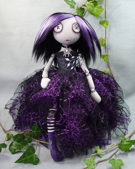 Gothic-style art doll with button eyes, purple hair and dress surrounded by green leaves on a light background