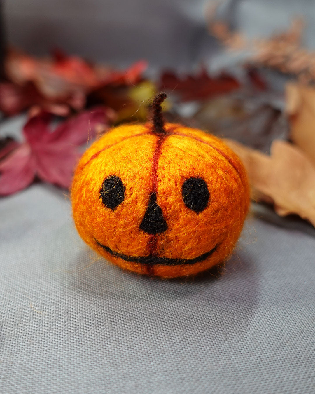 Small orange felt pumpkin with black face on a gray surface with autumn leaves in the background