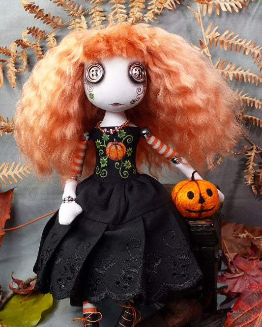 Doll with orange hair and black dress holding a small pumpkin, surrounded by autumn leaves.