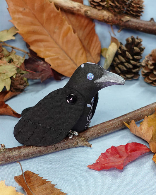 Textile crow on a branch with autumn leaves and pinecones in the background