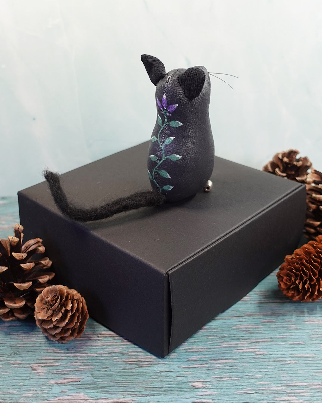 Black cat figurine with hand painted flower design on a black box with pine cones around, against a light blue background
