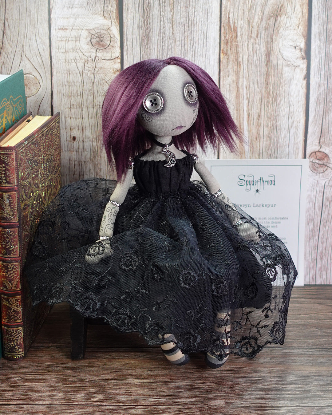 Gothic-style doll with purple hair and a black lace dress sitting next to a book on a wooden surface.