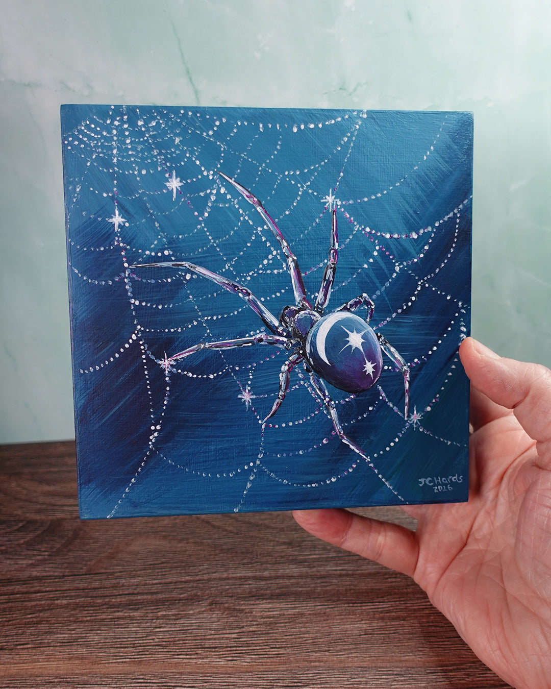 artist's hand holds a small square painting of a spider with moon and stars on its back in glistening web in blues and purples